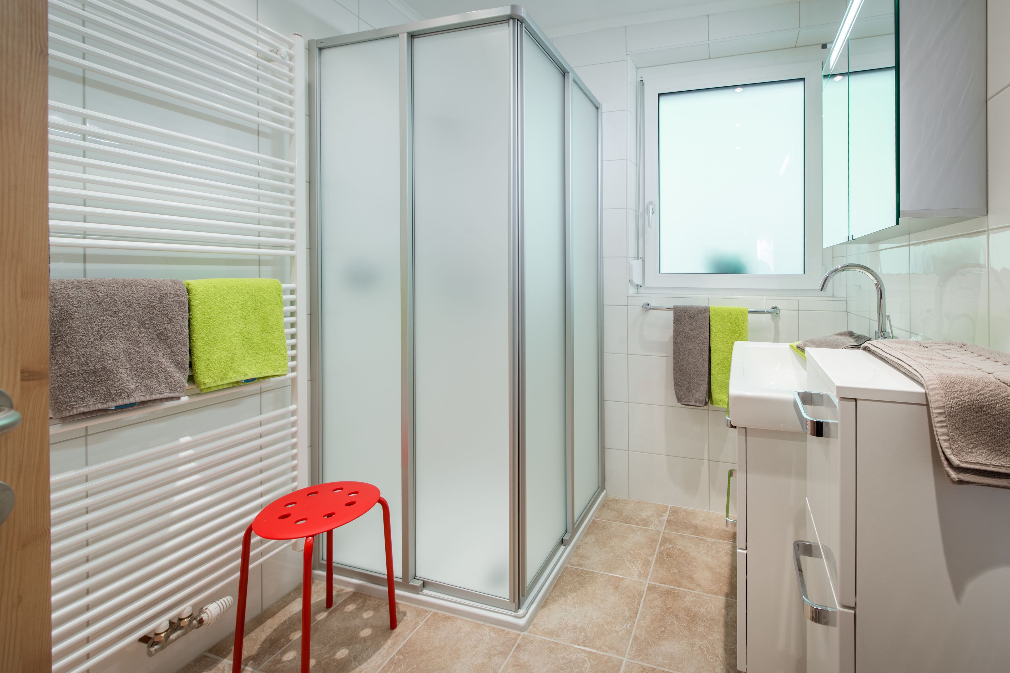 Ein modernes Badezimmer mit einer Duschkabine aus Glas, einem roten Hocker und einem Waschbecken mit Spiegel darüber.