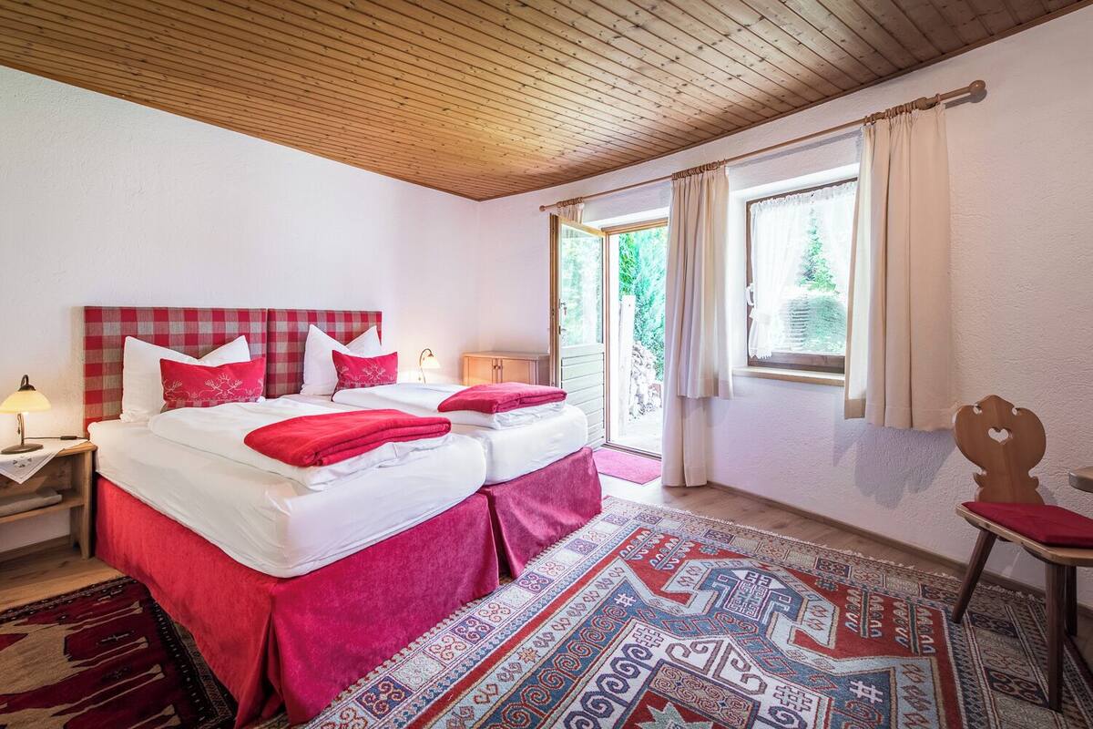 Ein Doppelbett mit roter und weißer Bettwäsche, ein Nachttisch mit Lampe, ein gemustertes Teppich und Blick auf Grünfläche.