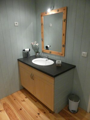 Un baño con tocador de madera, un lavabo blanco, un espejo con marco de madera, un secador de pelo y un cubo de basura.