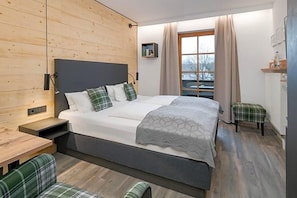 A modern bedroom with a large bed, a wooden headboard, a bedside table, a bench, and a window with curtains. - Hotel Neuer Am See (Prien am Chiemsee)