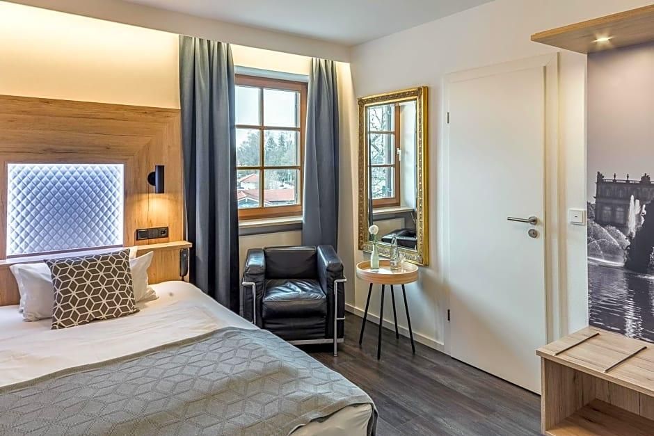 Ein Hotelzimmer mit Bett, Sessel, kleinem Tisch, Fenster mit Vorhängen, Tür und einem Wandspiegel.