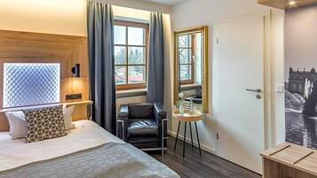 Ein Hotelzimmer mit Bett, Sessel, kleinem Tisch, Fenster mit Vorhängen, Tür und einem Wandspiegel.