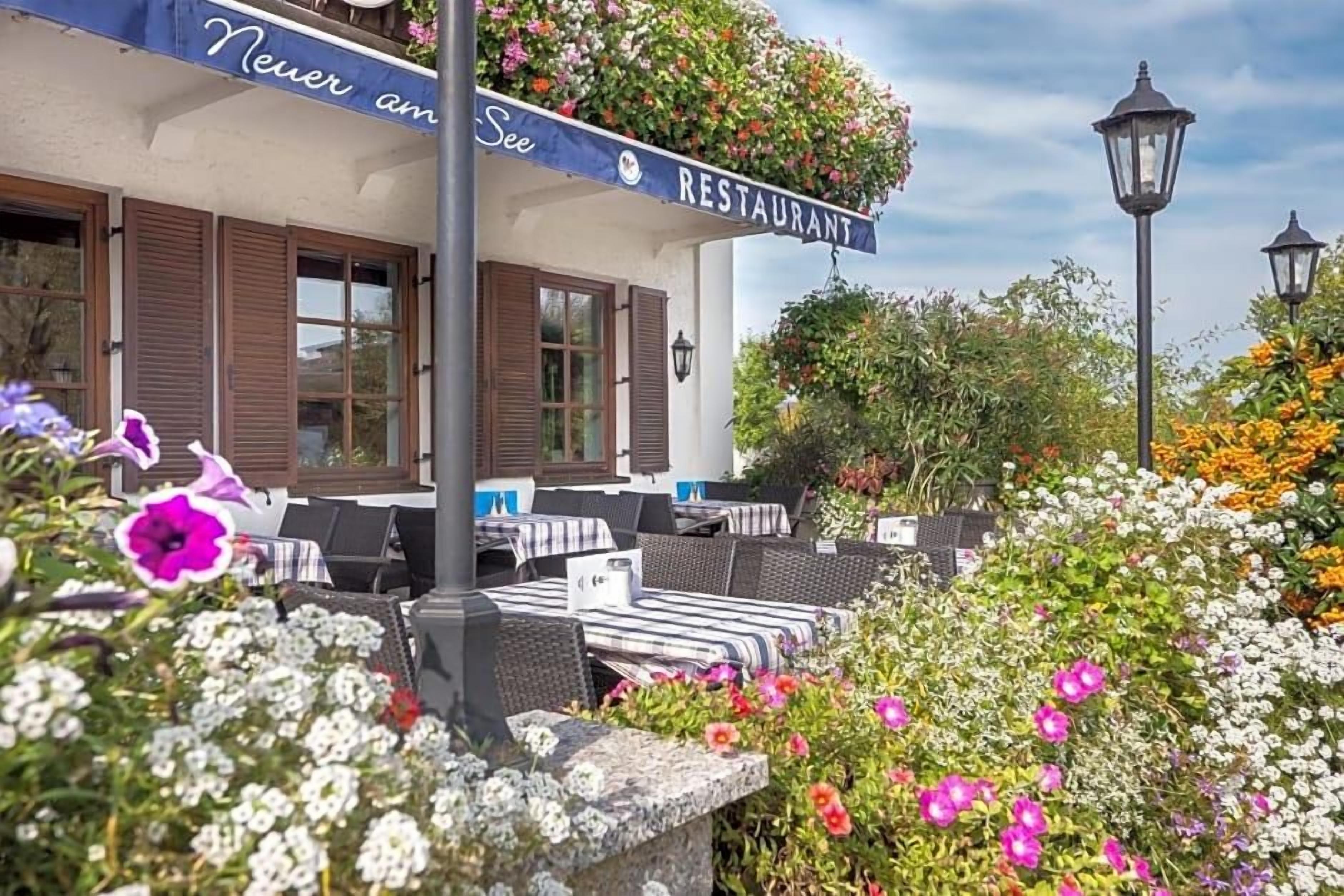 An outdoor seating area with tables and chairs, a sign reading 'Never am See Restaurant,' and a variety of flowers and plants.