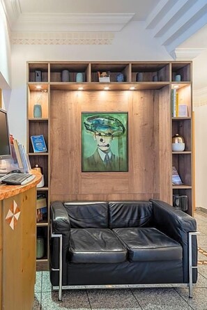 Une pièce avec un canapé en cuir noir, des étagères en bois où sont disposés divers objets, et un tableau encadré accroché au mur.