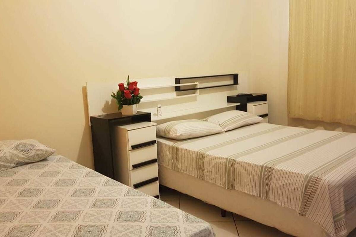 Ein Schlafzimmer mit einem Bett, zwei Nachttischen, einer Lampe, einer Vase mit Blumen und einem Fenster mit Vorhängen.