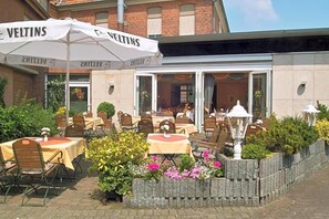 An outdoor seating area with tables and chairs, a brick building in the background, and a white umbrella with 'VELTINS' branding.