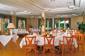 A dining area with multiple tables set for guests, featuring wooden chairs and white tablecloths.