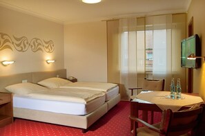A hotel room with a bed, bedside tables, a TV, a window with curtains, and a small table with bottles.