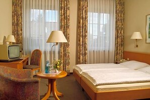 A hotel room with a bed, a chair, a small table with a lamp, a television, and curtains.