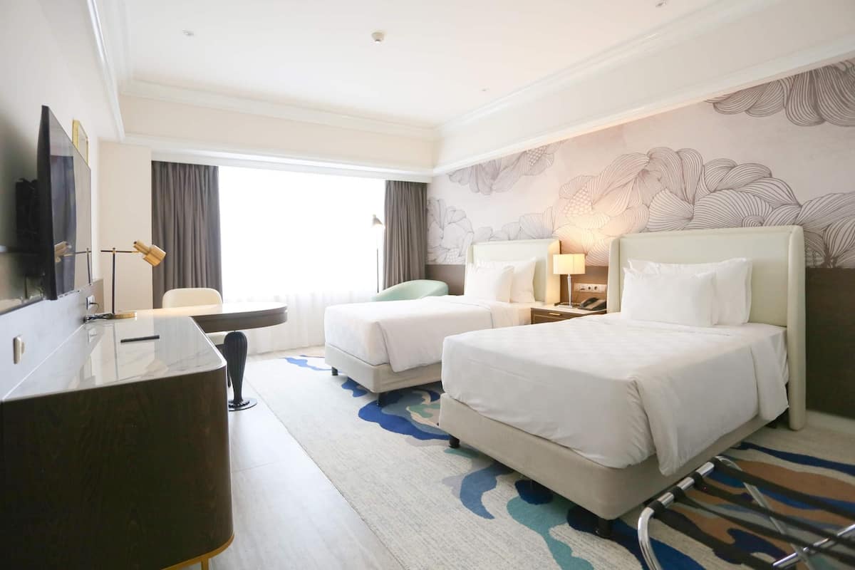 A hotel room with two beds, a desk, a TV, and a large window with curtains.