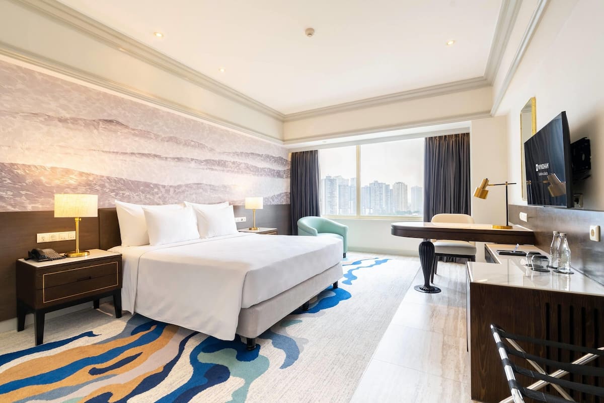 A hotel room with a large bed, a desk, a chair, a nightstand, a lamp, and a view of the city.