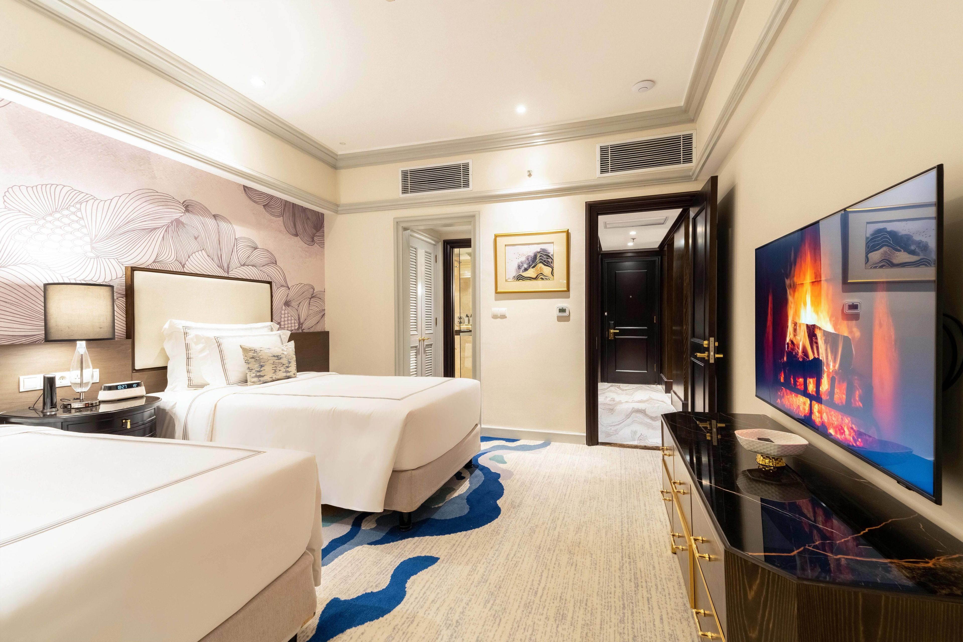 A hotel room with two beds, a TV, a desk with a lamp, and a decorative wall.