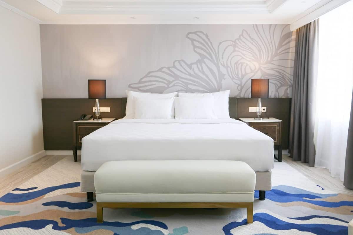 A hotel room with a large bed, two bedside tables with lamps, a bench at the foot of the bed, and a patterned carpet.