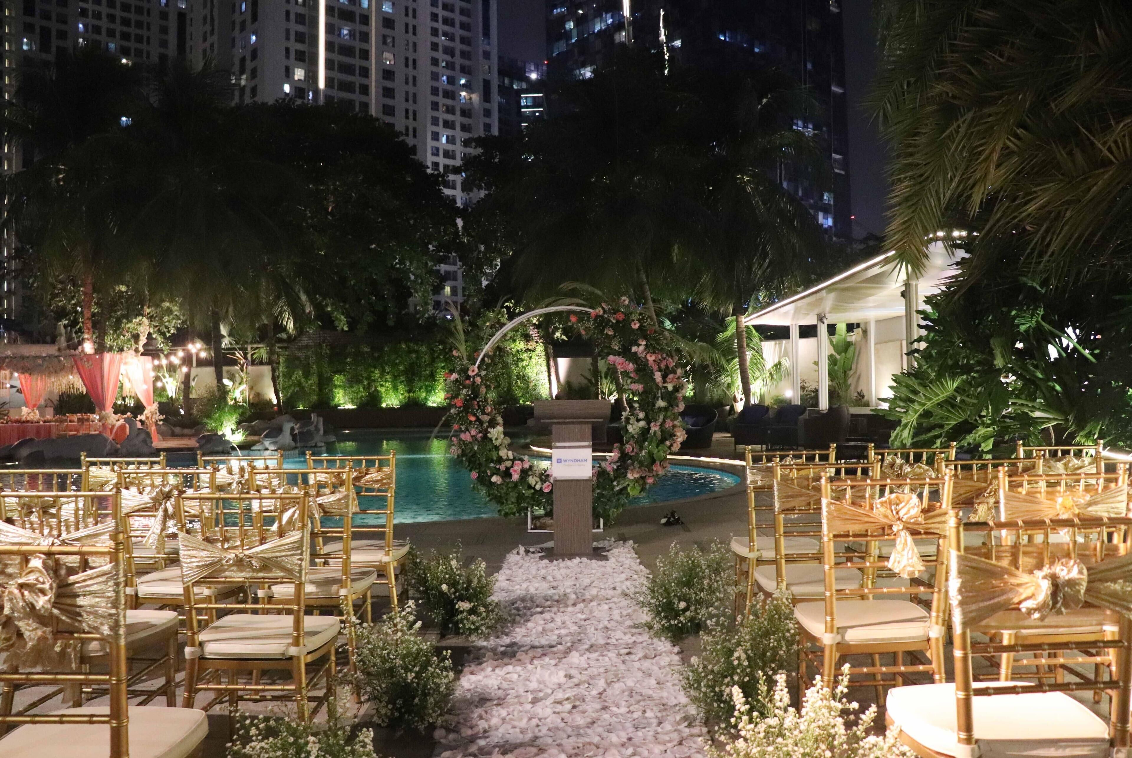 Un salón de bodas al aire libre con hileras de sillas doradas, un arco floral y una piscina.