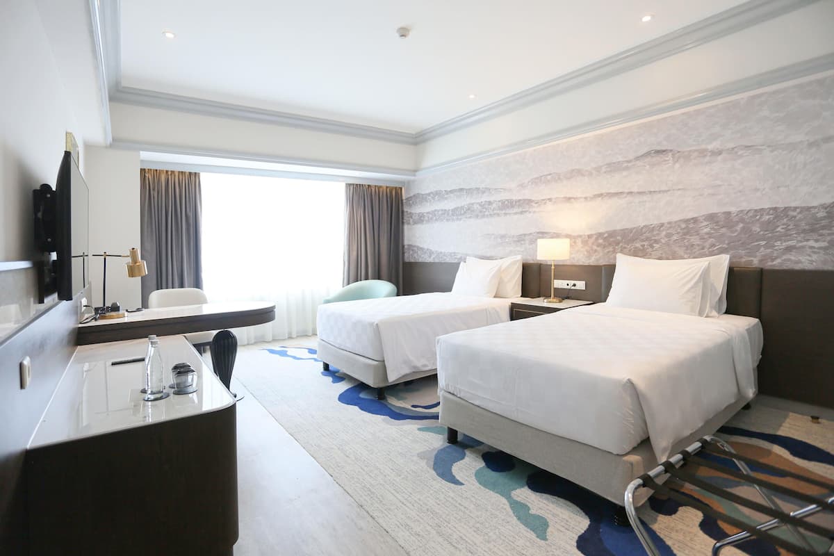 A hotel room with two beds, a desk, a chair, a TV, and a window with curtains.