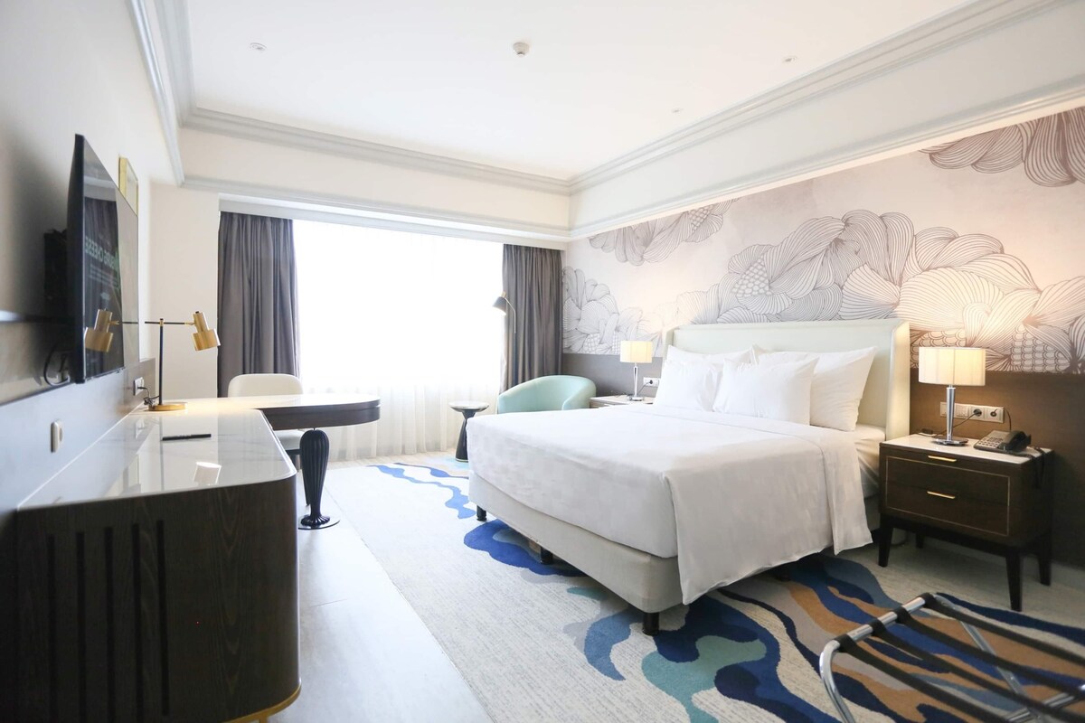 A hotel room with a bed, bedside table, lamp, desk, chair, and a large window with curtains.