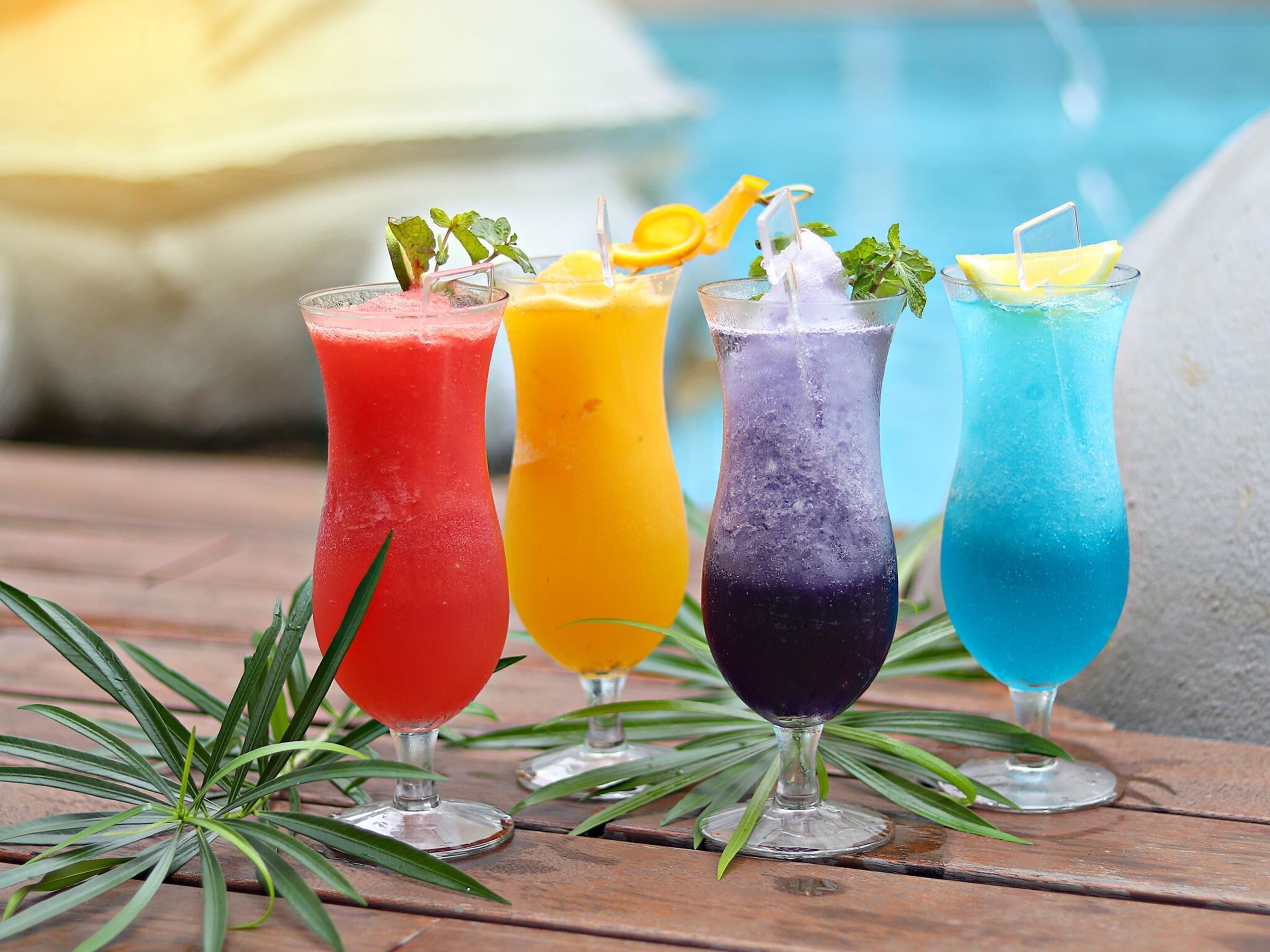 Four colorful drinks in glasses with garnishes, placed on a wooden surface with green plants.