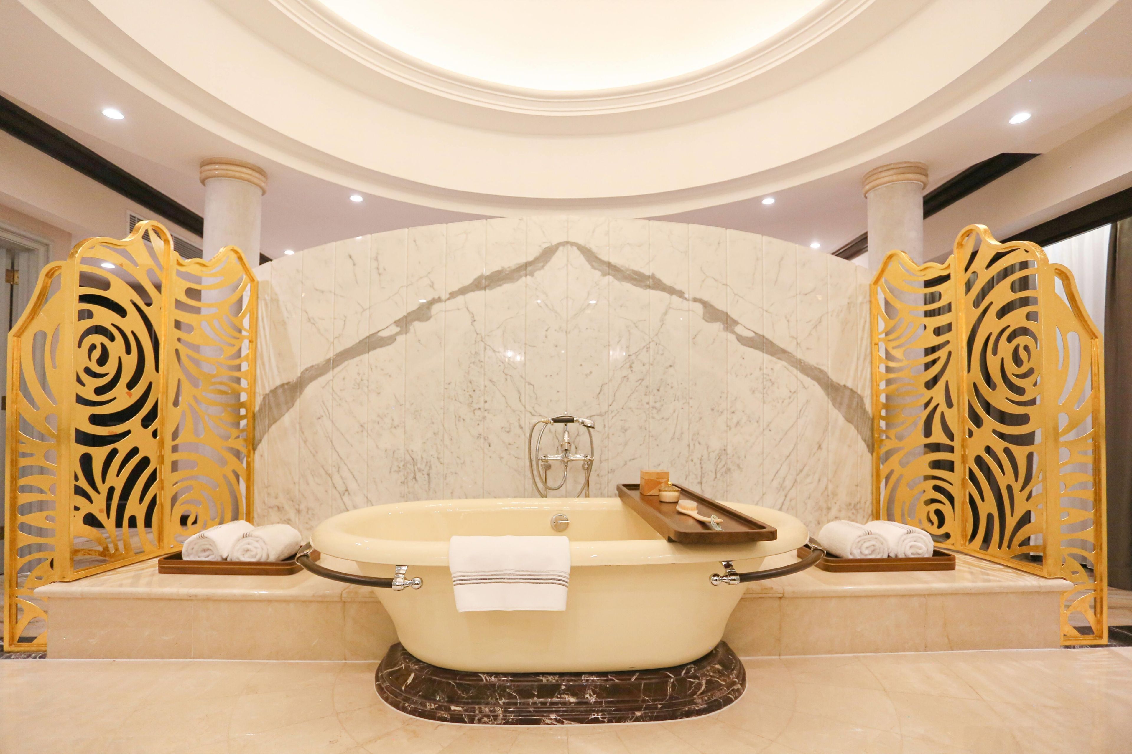 A large freestanding bathtub with a marble surround, two towel racks with towels, and decorative golden railings.