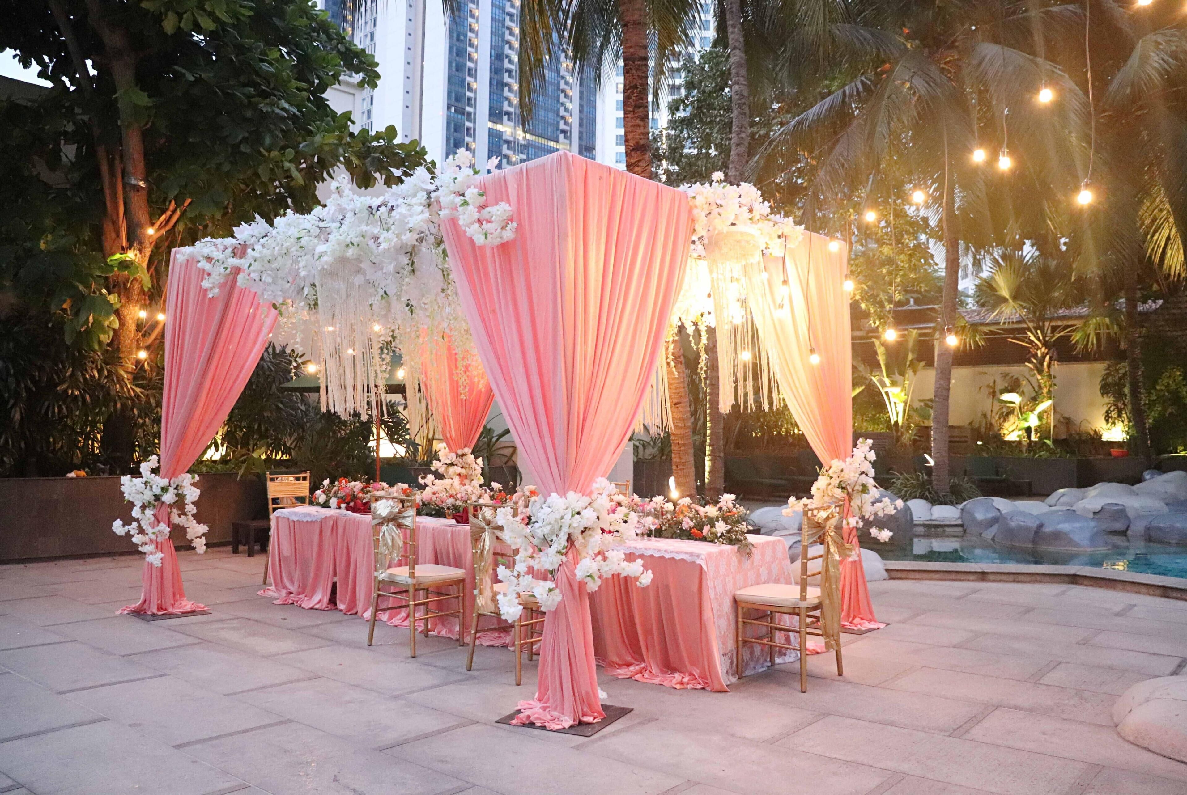 An outdoor event space with tables set for a reception, draped in pink and white fabric, and decorated with floral arrangements.