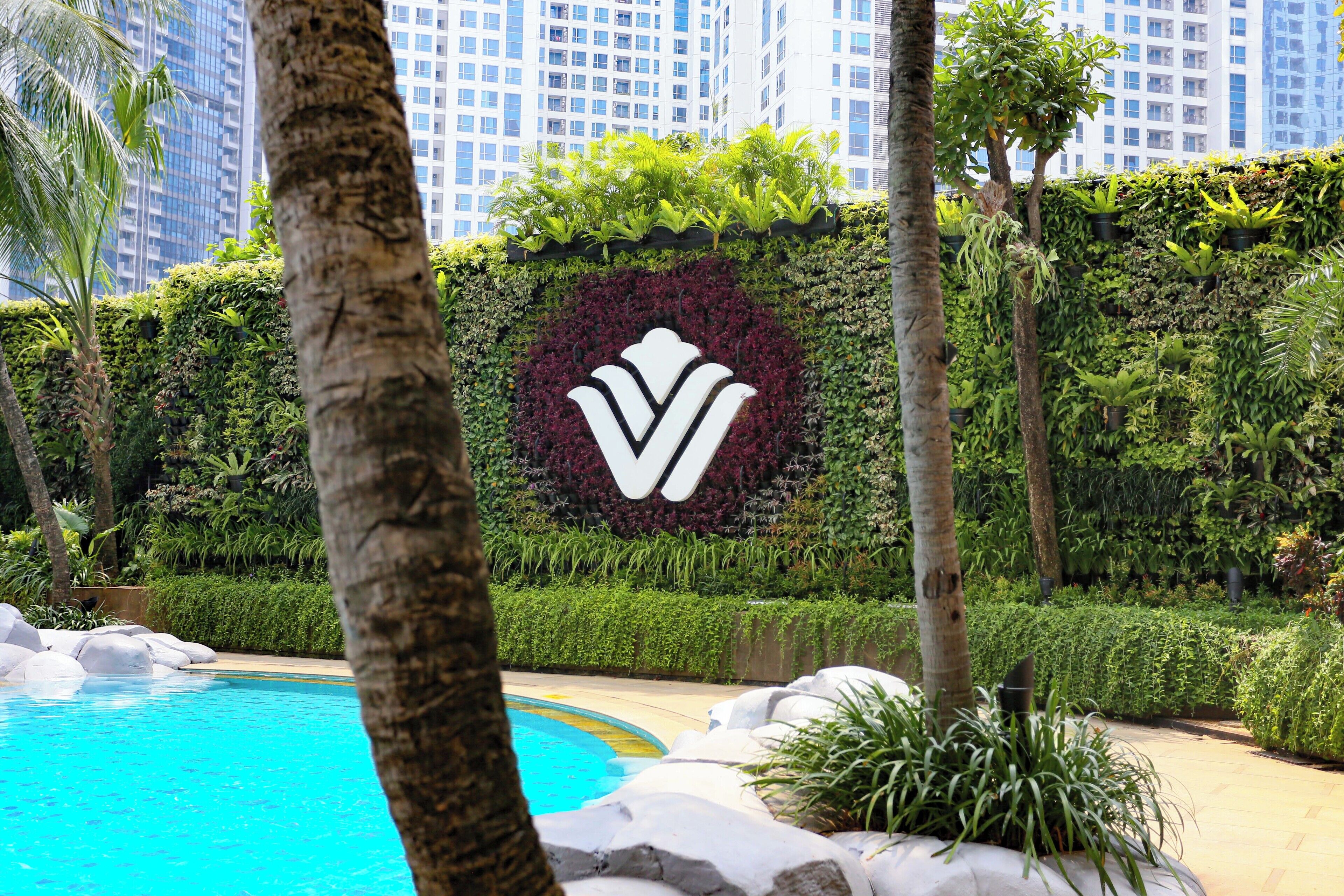 Un área ajardinada con piscina, un muro decorativo con un logotipo y edificios altos al fondo.