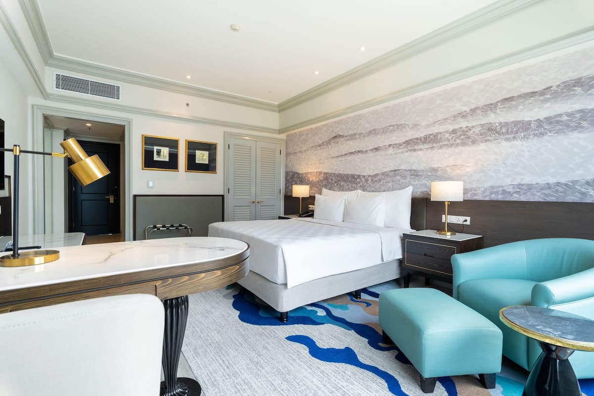 A hotel room with a bed, a desk, a chair, a round table, and a wall with a textured mural.