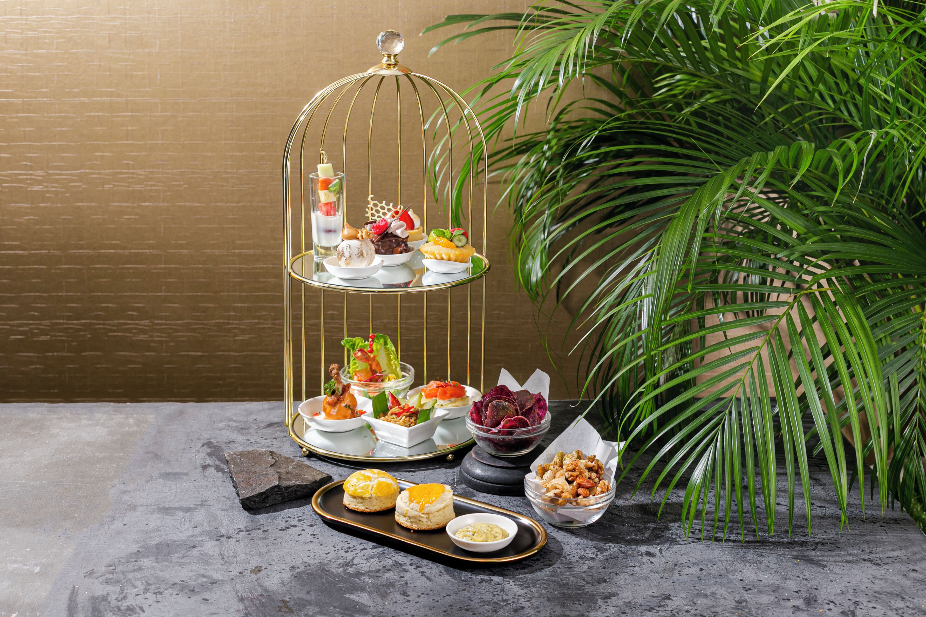 Une présentation de desserts à plusieurs niveaux, avec divers bonbons et pâtisseries, accompagnée d’un plateau de biscuits et d’un bol de noix, le tout mis en valeur par un mur de briques et une grande plante verte en arrière-plan.