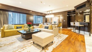 A spacious living room with a yellow sofa, a white ottoman, and a coffee table. There is a kitchen area with a bar and two bar stools. The room has large windows with a city view. - Wyndham Casablanca Jakarta (Jakarta)