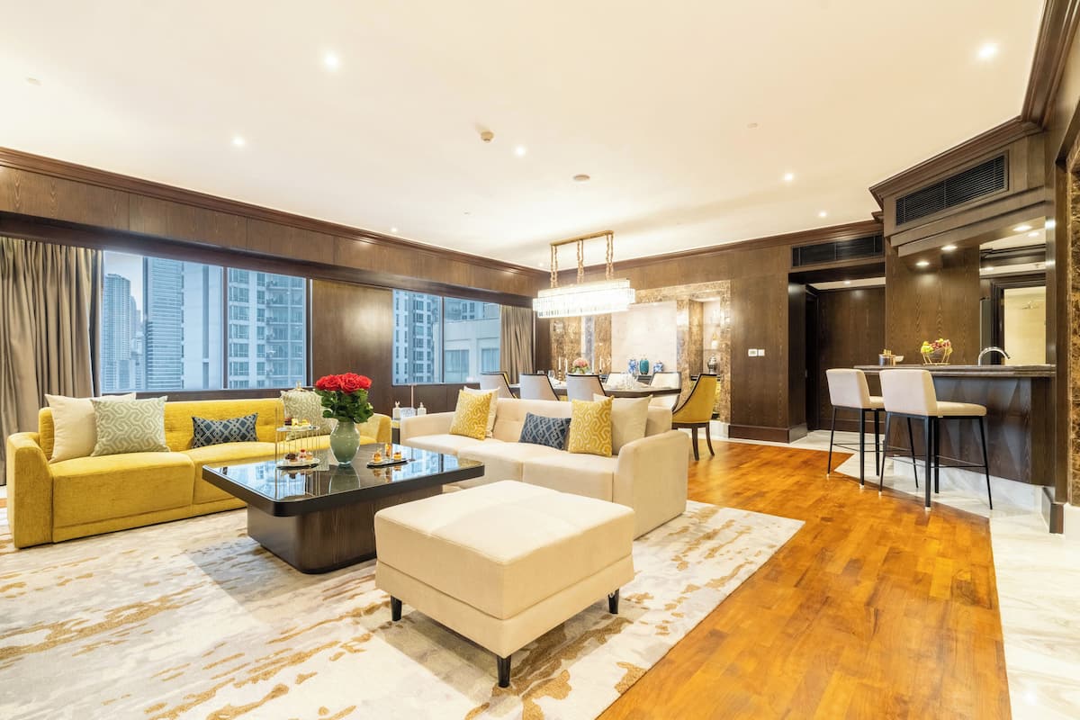 A spacious living room with a yellow sofa, a white ottoman, and a coffee table. There is a kitchen area with a bar and two bar stools. The room has large windows with a city view.
