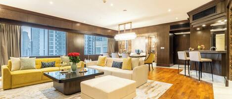 A spacious living room with a yellow sofa, a white ottoman, and a coffee table. There is a kitchen area with a bar and two bar stools. The room has large windows with a city view.