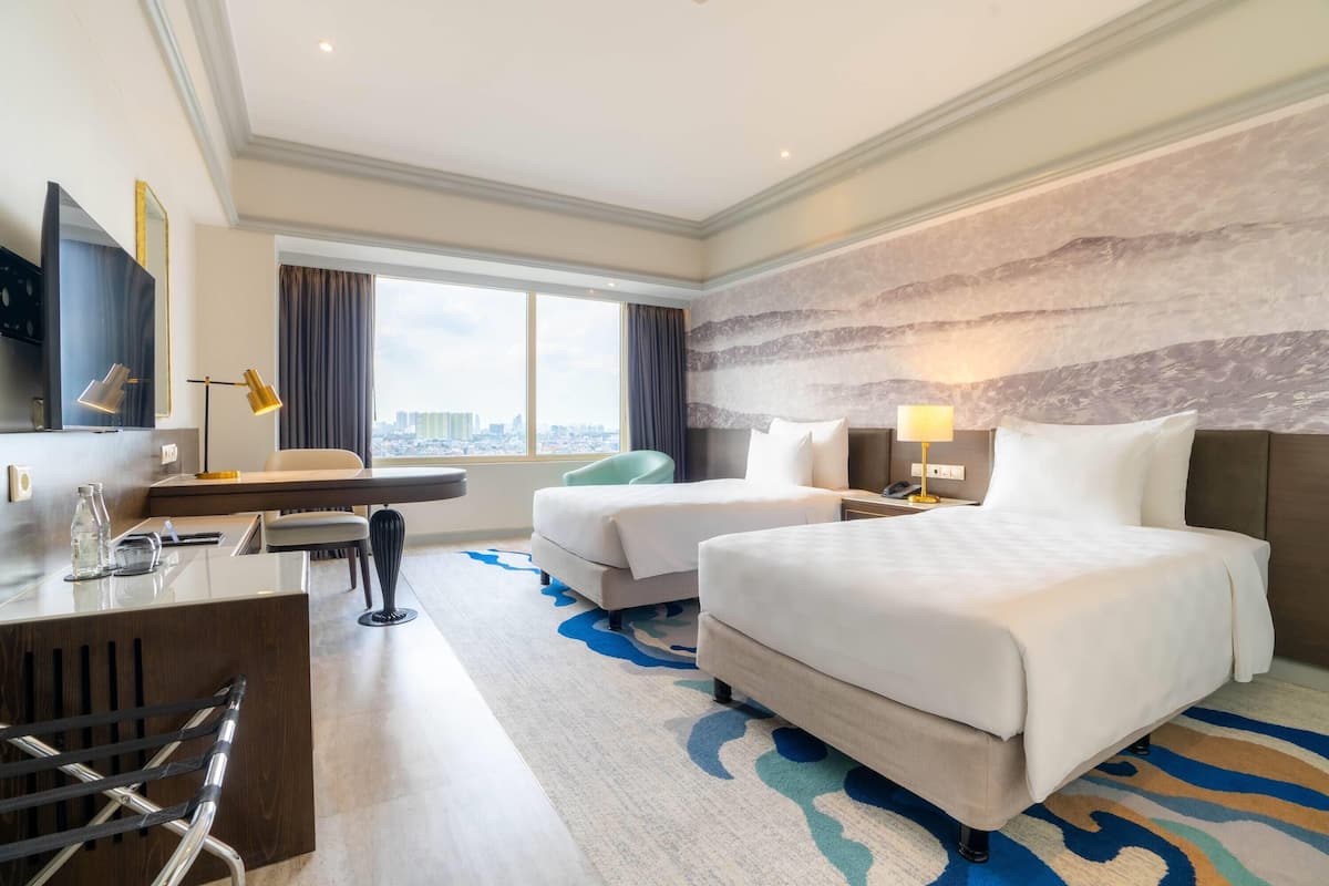A hotel room with two beds, a desk, a chair, a TV, and a large window with a city view.