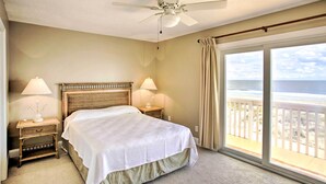Un dormitorio con cama, mesitas de noche, ventilador de techo y vistas a la playa a través de amplias ventanas.