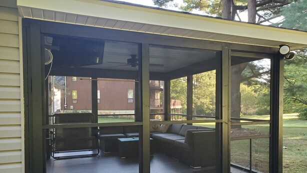 Een overdekt terras met een bank en een tafel, met uitzicht op een achtertuin met gazon en een gebouw op de achtergrond.