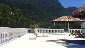 A pool area with a view of a mountain, a thatched-roof structure, and a white fence. - New Year's Eve 2026 - house + housekeeping services. Convenience and comfort (São Sebastião)