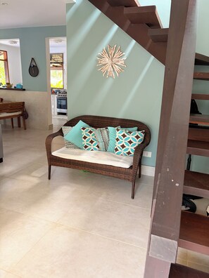 A wicker sofa with patterned cushions, a kitchen in the background, and a staircase.