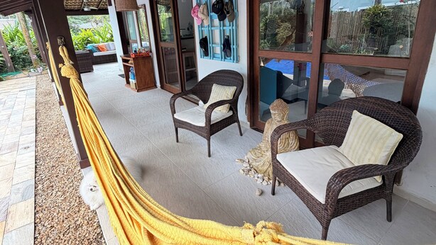 A covered outdoor seating area with wicker furniture, a yellow hammock, and a Buddha statue. - New Year's Eve 2026 - house + housekeeping services. Convenience and comfort (São Sebastião)