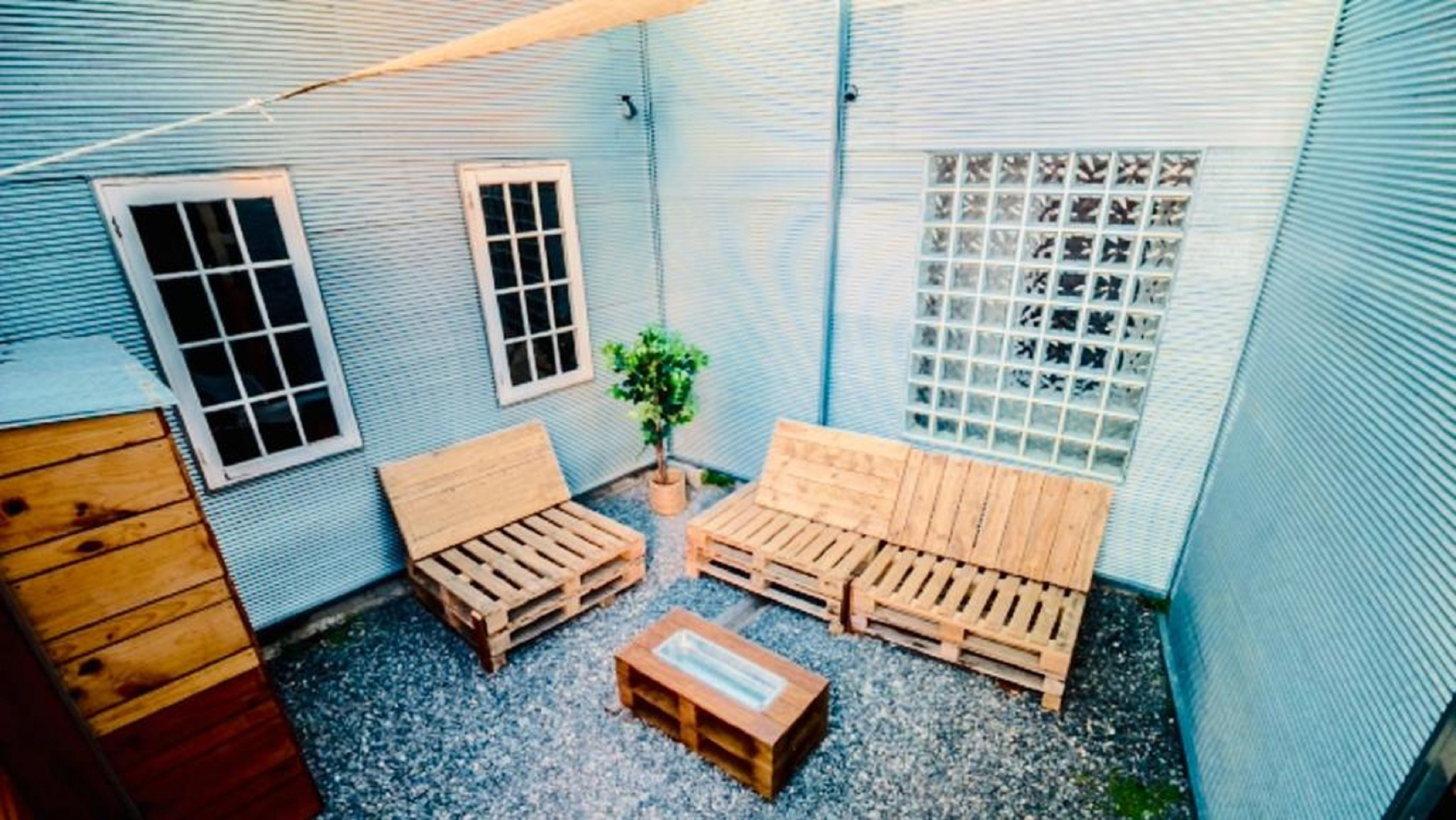 A small, enclosed outdoor space with a blue corrugated metal wall, two windows, a wooden bench, a small table, and a potted plant.