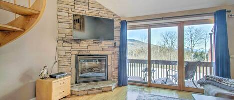 A living room with a stone fireplace, a wooden staircase, a flat-screen TV, and a large sliding glass door leading to an outdoor deck.