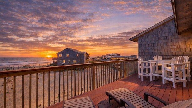 En träveranda med Adirondack-stolar och ett bord, med utsikt över en strand och ett hus.