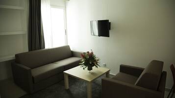 Una sala de estar con un sofá, una mesa de centro con un florero, un televisor montado en la pared y una ventana con cortinas.