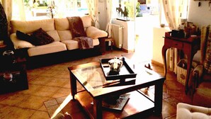 Ein Wohnzimmer mit einer Couch, einem Couchtisch und einem Sessel. Es gibt ein Fenster mit Vorhängen, eine Heizung und einen kleinen Tisch mit Dekoration.