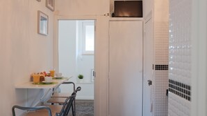 Un pasillo estrecho que conduce a un comedor con una mesa para dos, una planta pequeña y una puerta blanca con un teléfono.