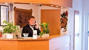 A reception area with a wooden counter, a receptionist, a potted plant, and a vase with flowers. - Rosenhof (Kerpen)