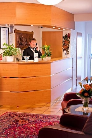 Un área de recepción con mostrador de madera, una recepcionista, una planta en maceta y un jarrón con flores.