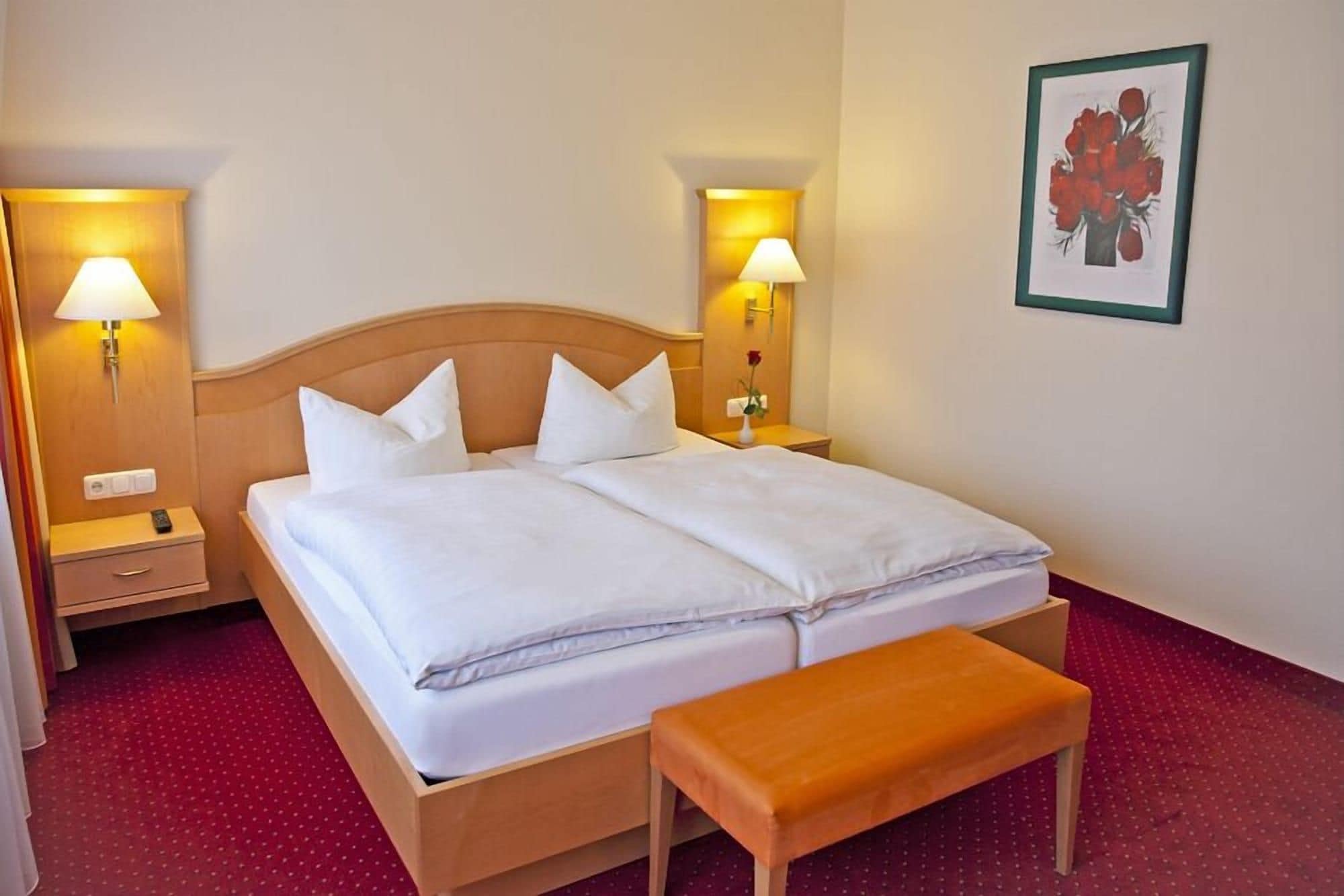 Ein Doppelbett mit weißen Leinen, zwei Nachttischlampen, ein Holz-Kopfteil, ein kleiner Nachttisch, ein roter Teppich und ein gerahmtes Bild an der Wand.