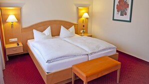A double bed with white linens, two bedside lamps, a wooden headboard, a small bedside table, a red carpet, and a framed picture on the wall. - Rosenhof (Kerpen)