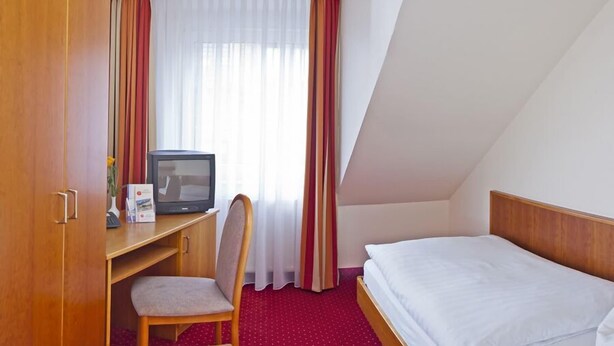 A hotel room with a single bed, a desk with a chair, a television, and a window with curtains. - Rosenhof (Kerpen)