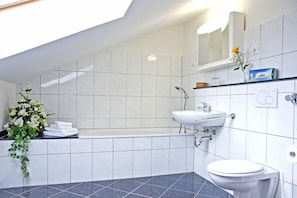 Un baño moderno con bañera, lavamanos e inodoro. Hay un espejo sobre el lavamanos y un arreglo floral a la izquierda.