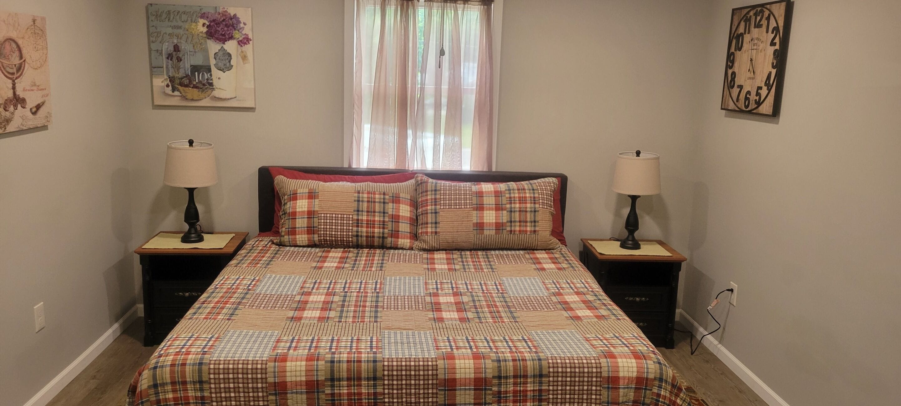 Un dormitorio con una colcha a cuadros, dos mesitas de noche con lámparas, un ventanal con cortinas de gasa y una obra de arte en la pared.