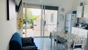 En kompakt opholdsstue med sofa, spisebord, stole, et tekøkken med mikroovn og en skydedør af glas til en udendørs terrasse.