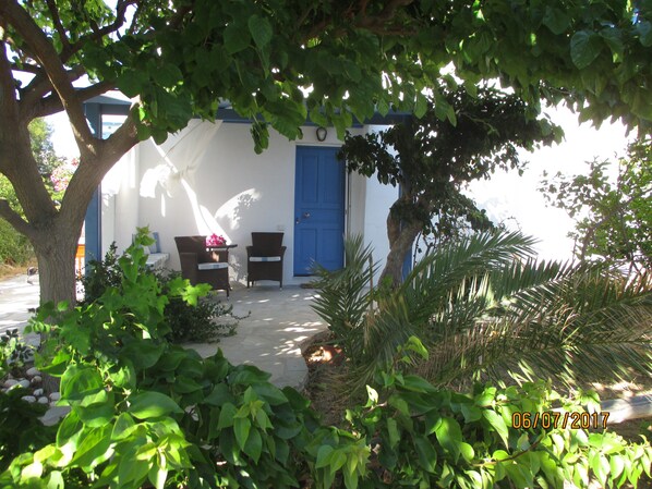 A covered outdoor seating area with two wicker chairs and a table, surrounded by lush greenery and a blue door. - ''Sea shell'' a jewel in plaka , Naxos (Plaka , Naxos)